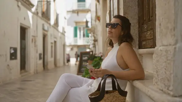 stock image A young hispanic woman uses her smartphone while relaxing in the picturesque white streets of locorotondo, puglia, italy.