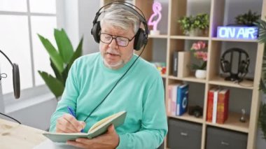 Radyo stüdyosunda kulaklık takan olgun bir adam yayın hazırlığı ya da podcast aktivitesini gösteren notlar yazıyor.