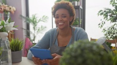 Çiçekçi dükkanında tablet kullanan neşeli bir Afro-Amerikalı kadın yemyeşil bitkilerle çevrili.