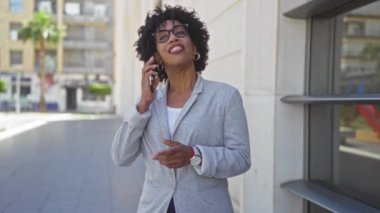 Kıvırcık saçlı, kendine güvenen Afro-Amerikalı kadın şehir sokağında cep telefonuyla konuşurken gri bir ceket ve gözlük takıyor ve neşe ve bağlantı kuruyor..