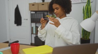Kıvırcık saçlı Afrikalı genç bir Amerikalı kadın ofis ortamında telefonuyla fotoğraf çekiyor. Etrafı kitaplarla ve ofis malzemeleriyle çevrili..