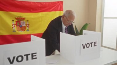 İspanya 'da kapalı bir oylama odasında kıdemli bir adam, büyük bir İspanyol bayrağıyla işaretlenmiş, bir seçim sürecine katılıyor,' oy 'işaretleriyle süslenmiş bir oy kabininde duruyor..