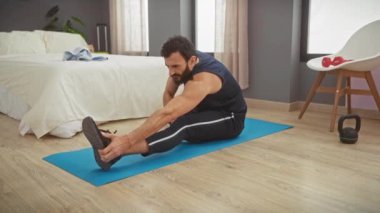 Orta yaşlı, sakallı bir adam, rahat bir yatak odasında mavi bir paspasa uzanıyor, spor yapıyor ve yaşam tarzını gösteriyor..