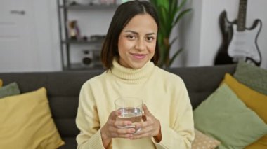 Gülümseyen genç bir kadın, arka planda gitarıyla rahat bir oturma odasında elinde bir bardak su tutuyor..
