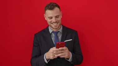 Gülümseyen, takım elbiseli genç İspanyol adam izole edilmiş kırmızı arka planda akıllı telefondan mesaj atıyor. Serin gülümsemesi ve ışıldayan dişleriyle mutluluk ve kendine güven yayıyor.
