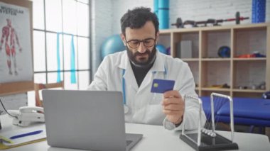 Gözlüklü sakallı bir doktor, fizyoterapi kliniğindeki bilgisayarında kredi kartını inceliyor..