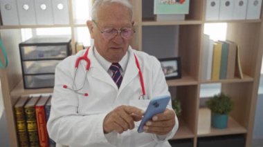 Beyaz önlük ve steteskop giyen yaşlı bir adam, arka planda kitaplıklar olan bir klinikte akıllı telefon kullanıyor..