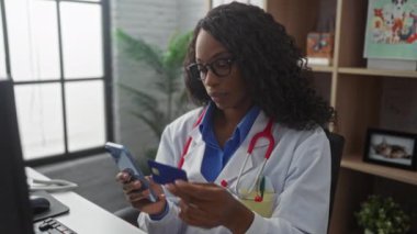 Afrikalı Amerikalı bir kadın doktor klinikte steteskop ve gözlük takarak kredi kartı ve cep telefonu kullanıyor..