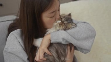 Genç Çinli bir kadın, evindeki rahat oturma odasında tüylü kedisini öpüyor ve kedi yavrusunu sevgiyle kucaklıyor..