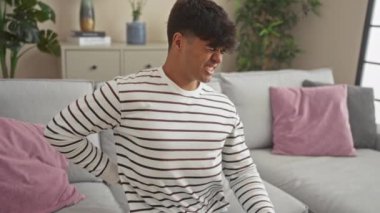 Çizgili gömlekli genç İspanyol adam, modern bir oturma odasındaki kanepede bitki ve dekoratif yastıklarla otururken sırt ağrısı çekiyor..