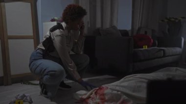 Afro-Amerikalı kadın olay yerini kan, telefon, eldiven, kanıt işaretleyiciler ve entrikayla inceliyor..