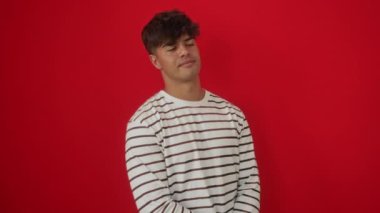 Üzerinde çizgili tişört olan İspanyol genç adam kameraya bakıyor ve gülümsüyor. Elinde çenesi, kolları bağlı, izole edilmiş kırmızı arka planda pozitif düşünüyor.