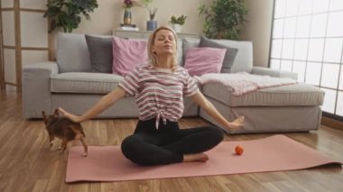 Sarışın genç bir kadın, Chihuahua 'sıyla yoga yapıyor rahat bir oturma odasında, içinde bitkiler, bir kanepe ve pembe bir spor paspas var..