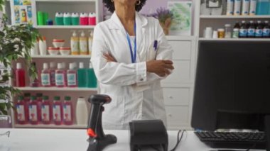 Kıvırcık saçlı, kendinden emin bir Afrikalı Amerikalı kadın eczacı kolları çaprazlanmış bir eczanede duruyor. Etrafı çeşitli şişe ve eczane malzemeleriyle çevrili..