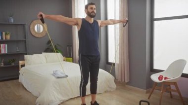 Sakallı adam, modern bir yatak odasında direniş bantlarıyla egzersiz yapıyor.