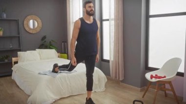 Atletik giyinen sakallı bir adam modern bir yatak odası ortamında esneme hareketleri sergiliyor..