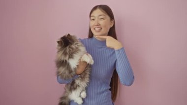 Genç Çinli kadın ve kedi el ve parmaklarıyla birbirlerini gösteriyorlar, mutlu ve gülümsüyorlar izole pembe arka planda