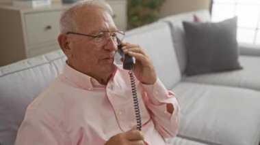 Pembe gömlekli kıdemli beyaz adam ev telefonu kullanıyor ve evdeki rahat oturma odasında gri bir kanepede oturuyor..