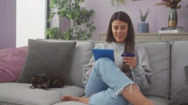 Tablet kullanan ve yanında Chihuahua olan bir kredi kartı tutan gülümseyen bir kadın.