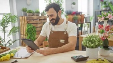 Sakallı adam çiçekçide tablet kullanıyor. Etrafı bitki ve buketlerle çevrili..
