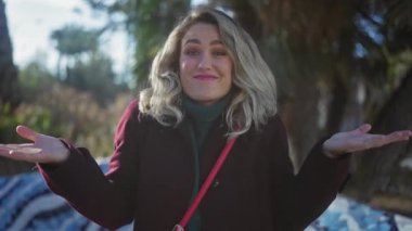 Sarışın genç bir kadın, şehir parkında paltosunu giymiş, ellerini kaldırarak dışarı çıktı.