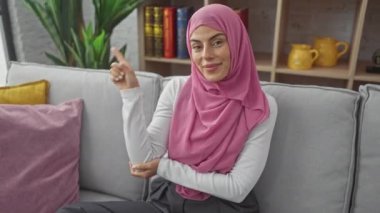 Neşeli, kendinden emin, tesettürlü, tesettürlü genç bir kadın, mutlu bir şekilde parmak ucunda, evde bir kanepede gülümserken.