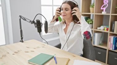 Kulaklıklı olgun bir kadın, podcast stüdyosunda mikrofona konuşuyor. Kapalı alanda profesyonellik ve kendine güven yayıyor..
