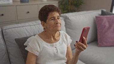 Kısa saçlı yaşlı İspanyol bir kadın evde oturma odasında otururken kırmızı bir akıllı telefondan video görüşmesi yapıyor.