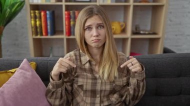 Genç sarışın kadın, üzgün ve mutsuz, somurtkan bir şekilde kanepede oturuyor, üzgün üzgün bir bakışla aşağıya bakıyor, depresif bir şekilde yön gösteriyor. Desmotivasyonun dokunaklı bir portresi..