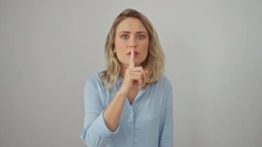 Çekici sarışın kadın rahat poz veriyor, tişört giyiyor, dudakları parmaklarıyla işaret ediyor. Genç yetişkinler, beyaz izole edilmiş arka planda gizli bir sessizlik sinyali veriyor. Sessiz zarafetin portresi.