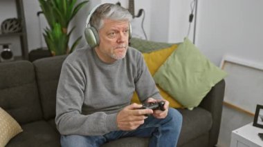 Oturma odasındaki kanepede video oyunu oynayan kulaklıklı olgun bir adam heyecan ve hayal kırıklığını ifade ediyor..