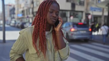 Güneşli bir şehir caddesinde yürürken cep telefonuyla konuşan Afrikalı Amerikalı kadın..