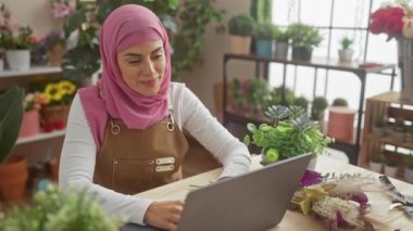 Başörtüsü takan gülümseyen bir kadın ev çiçekçisinin evinde dizüstü bilgisayarda çalışıyor. Başarılı ve neşeli görünüyor..