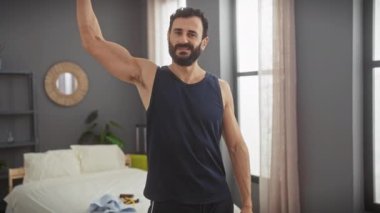 Orta yaşlı, sakallı bir adam modern yatak odasının içinde kaslarını gösteriyor.