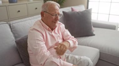 Pembe tişörtlü yaşlı bir adam, gri bir koltukta oturur. Kollarını kavuşturur, rahat bir oturma odasında..