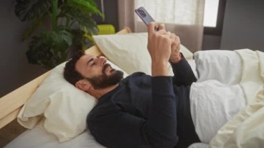 Yakışıklı sakallı adam yatak odasının içinde akıllı telefon kullanıyor.
