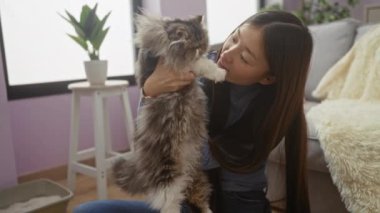 İçerde rahat bir ortamda genç bir kadın arka planda bir bitkiyle pofuduk yavrusunu oturma odasında tutar ve öper..