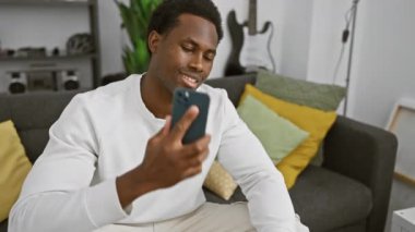 Neşeli bir adam gündelik kıyafetlerle akıllı bir telefonla konuşuyor, modern bir oturma odasındaki koltukta rahatça oturuyor..