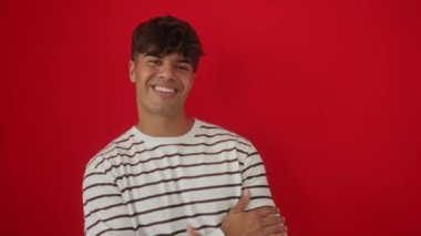 Çizgili tişört giyen, gülümseyen, gülümseyen ve baş parmağıyla yan tarafı gösteren İspanyol genç bir adam. izole edilmiş kırmızı arkaplan