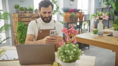 Çiçekçi dükkanı işleten sakallı bir adam telefonunu canlı bitkilerin ve dizüstü bilgisayarın arasında kontrol ediyor..