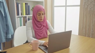 Pembe tesettürlü odaklanmış bir kadın evde kahvesini yudumlarken dizüstü bilgisayarla çalışıyor.