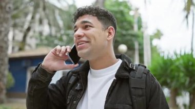 Kıvırcık saçlı ve sırt çantalı gülümseyen genç adam ağaçlarla dolu bir caddede akıllı telefondan konuşuyor..