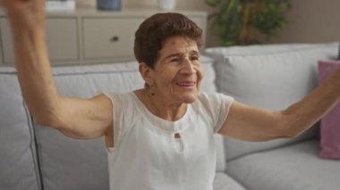 Neşeli, yaşlı bir kadın sıcak bir oturma odasında neşeli bir ifadeyle kutlama yapıyor ve kollarını kaldırıyor, beyaz bir bluz giyiyor ve modern bir arka plana karşı rahat bir kanepeye oturuyor..