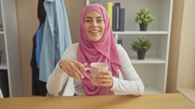 Başörtüsü takan gülümseyen kadın kitap rafı arka planında rahat bir evde bardak tutuyor..