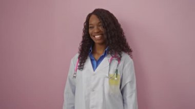Doktor, üniformalı genç Afro-Amerikan kadın kendine güvenen bir gülümseme ve gülen bir ifadeyle, soyutlanmış pembe arka planda, başka tarafa bakıyor, doğal ve mutlu.