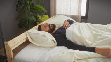 Orta yaşlı, sakallı bir adam yatakta geriniyor, sabah rutini ev yatak odası düzeninde..