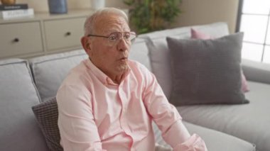 Oturma odasındaki kanepede oturan gri saçlı yaşlı bir adam, gözlük ve pembe gömlek giyerken çeşitli duygular sergiliyor..