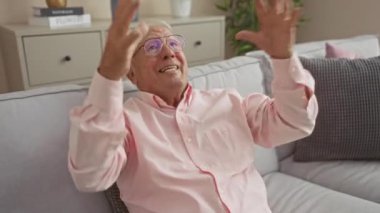 Gözlüklü, pembe tişörtlü, rahat bir oturma odasında kanepe ve iç mekan bitkileriyle heyecanını ifade eden yaşlı beyaz bir adam..