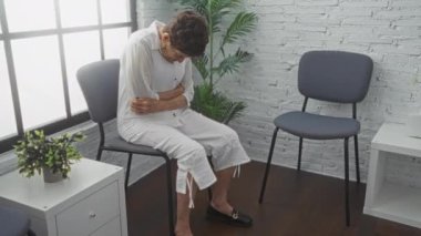 Yaşlı bir İspanyol kadın, gri sandalyeli ve yeşillikli modern bir bekleme odasında otururken karnını ağrıtıyor. Bu da kapalı bir tıbbi ya da klinik çevreye işaret ediyor..