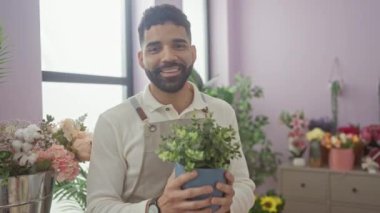 Sakallı, gülümseyen genç bir adam parlak, renkli bir çiçekçinin içindeki saksı bitkisini koklamaktan zevk alır..